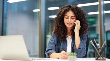 Kendine güvenen genç iş kadını telefonda konuşuyor ve modern ofiste notlar alıyor. Masasında çalışan kıvırcık saçlı kadın..