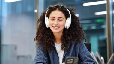 Güzel, neşeli esmer kadın modern bir ofiste kulaklıkla müzik dinliyor. Mutlu ve gülümsüyor..