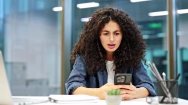 Sinirli iş kadını akıllı telefondan kötü haberler okuyor. Stresli, kıvırcık saçlı, günlük kıyafetler giyen, modern ofiste telefon kullanan bir kadın..