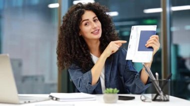 Kıvırcık saçlı iş kadını, ofiste kamerayla konuşurken pasaportunu ve uçak biletini gösteriyor..