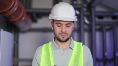 Kendine güvenen sakallı, kasklı ve üniformalı fabrika işçisi kazan dairesinde kameraya gülümsüyor..