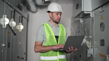Elektrik kontrol odasında dizüstü bilgisayarı olan kendinden emin bir elektrik mühendisi. Kontrol paneli olan elektrik santrali. Enerji uzmanı güç kaynağını inceliyor. Elektrik santralinde endüstri mühendisi.