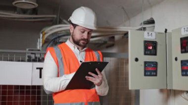 Beyaz şapka ve turuncu yelek giyen Kafkas elektrik mühendisi bir sunucu odasındaki elektrik santralini veya elektrik kontrol panelini inceler. Sunucu odasındaki endüstriyel elektrik kontrol paneli.