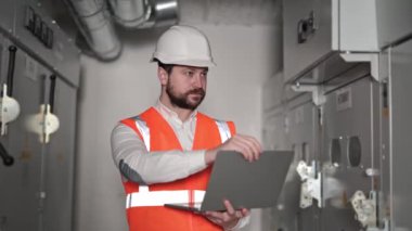 Dizüstü bilgisayarla çalışan endüstriyel elektrikçi, Elektrik Santralini Denetliyor. Sakallı Orange Yelekli Endüstriyel Mühendis Sunucu odasındaki Elektrik Kontrol Panelinde çalışıyor..