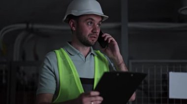 Üniformalı ve kasklı ciddi bir adam telefonda konuşurken not alıyor. Endüstriyel bina içi. Yanıp sönen kırmızı ışık. Sistem hatası, tehlike