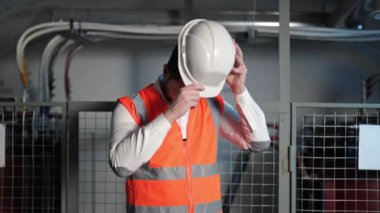 Kafkas elektrik mühendisi beyaz koruyucu başlık takıp el sallıyor. Sakallı, ciddi, beyaz gömlekli ve parlak turuncu çizgili bir adam..