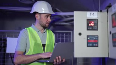 Ciddi elektrik mühendisi dizüstü bilgisayarla elektrik kontrol panelinin yanında çalışır. Santralin yanındaki bilgisayar klavyesinde sabit şapkalı ve üniformalı odaklanmış bir adam..