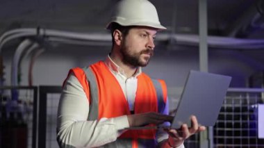 Elektrik santralinde dizüstü bilgisayarı olan bir elektrik mühendisi. Turuncu yelek giyen ve dizüstü bilgisayarında beyaz şapka takan adam yoğun bir şekilde işe odaklanmış. Arka planda elektrik kontrol paneli ve borular.