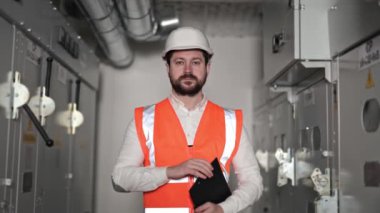 Ciddi bir elektrik mühendisi, sakalı, turuncu güvenlik yeleği ve beyaz kaskı olan bir klasör elektrik santralleri ve elektrik kontrol panelleri arasında duruyor..