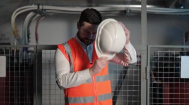 Sakallı elektrik mühendisi beyaz kaskı takıyor. Arka planda elektrik kontrol panelleri ve santralleri olan endüstriyel ortam.