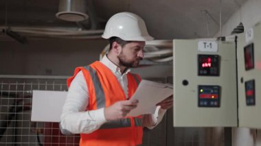 Turuncu yelek ve beyaz başlıklı ciddi bir elektrik mühendisi belgelerini tutuyor ve bir elektrik kontrol panelinin verilerini kontrol ediyor.