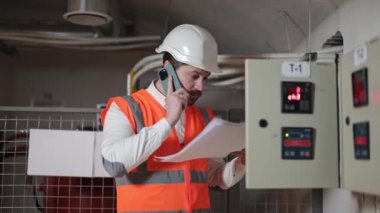 Turuncu yelek ve beyaz kasklı ciddi bir elektrik mühendisi telefonda dökümanları tartışıyor. Sakallı adam mobil cihaz kullanıyor, teknik odada belgeler okuyor..