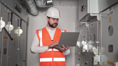 Profesyonel elektrik mühendisi, dizüstü bilgisayarla elektrik kontrol panelinin yanında çalışır. Sakallı adam turuncu yelek ve beyaz kasklı tipler dizüstü bilgisayar klavyesinde ve kameraya bakıyor..