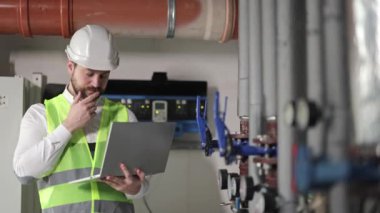 Beyaz kasklı ve floresan yeşili yelekli sakallı adam elektrik santralinin kazan dairesinde boru ve göstergelerin yanında dizüstü bilgisayarla çalışıyor..