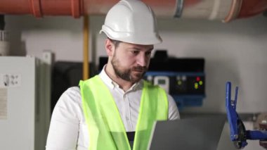 Kazan dairesindeki sakallı işçi ısıtma sistemini incelemek için dizüstü bilgisayar kullanıyor. Endüstriyel çevre. Elektrik üretim fabrikası işçisi.