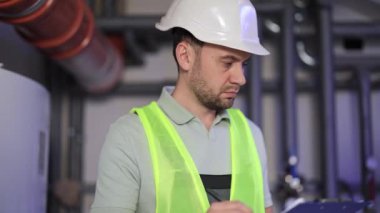 Kazan dairesinde güvenlik kaskı ve yeleği takan ciddi bir erkek mühendis, panoya yazı yazıyor. Borular ve ekipmanlarla endüstri ortamı.