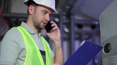 Kazan dairesindeki ciddi adam ısıtma sistemi ölçümlerini kontrol ediyor. Kask ve üniforma konusunda endüstriyel uzman telefonda konuşuyor, belgeyi okuyor.