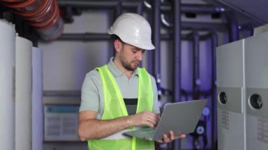 Kazan dairesindeki kendine güvenen erkek mühendis dizüstü bilgisayarı kullanıyor ve büyük kazanlar, borular ve ısıtma sistemi ekipmanlarının yanında dururken onu kapatıyor..