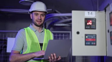 Elektrik mühendisi, elektrik santralinin yanında dizüstü bilgisayar kullanır. Kask ve güvenlik yeleği giyen profesyonel bir adam ekipmanları kontrol ediyor. Dijital kontrol paneli olan güvenli sunucu odası.