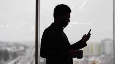 Kahve içen ve pencerenin yanında akıllı telefon kullanan düşünceli bir Afrikalı işadamı silueti. Şehir manzaralı modern ofis binası.