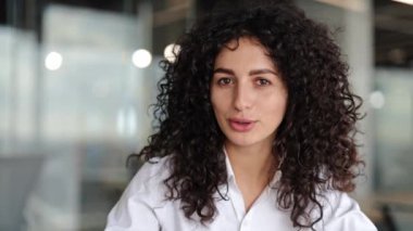 Kendine güvenen kıvırcık saçlı genç iş kadını modern bir ofiste kameraya gülümsüyor. Profesyonel ve dost canlısı görünüyor..