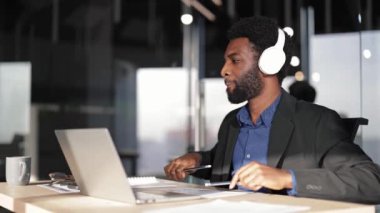 Modern ofis masasında çalışan odaklanmış bir iş adamı. Takım elbiseli şık siyah erkek müzik dinler ve dizüstü bilgisayarla çalışır. Şirket çalışanı belgeleri araştırıyor..