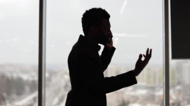 Modern ofisteki büyük pencereli iş adamının silueti telefonda konuşuyor. Kendine güvenen girişimci akıllı telefondan bir anlaşma yapar. Ciddi iş tartışması.