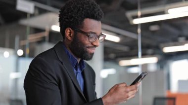 Gülümseyen genç Afrikalı işadamı gözlük takıyor ve modern ofiste cep telefonu kullanıyor. Akıllı telefonu iş ve iletişim için kullanan kendine güvenen bir adam..