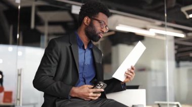 Odaklanmış işadamı, akıllı telefon kullanarak belgelerden gelen verileri doğruluyor. Genç Afrikalı erkek girişimci modern ofiste çalışıyor..