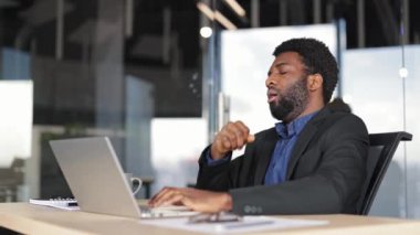 Modern ofisteki iş yerinde esnemekten bıkmış bir işadamı. Yorgun düşmüş Afrikalı Amerikalı erkek işçi uykulu hissediyor, dizüstü bilgisayarla çalışıyor..