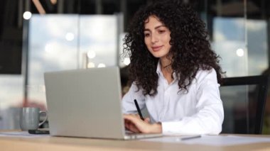 Modern ofiste dizüstü bilgisayarda çalışan genç kıvırcık iş kadını. İşyerinde bilgisayar kullanan beyaz gömlekli kadın çalışana odaklan..