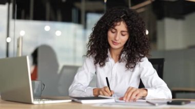 Modern ofiste çalışan kıvırcık saçlı, odaklanmış bir iş kadını. Online toplantıya katılırken deftere yazıyor..