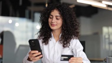 Modern ofiste online alışveriş yapmak için akıllı telefon ve kredi kartı kullanan kendine güvenen genç bir iş kadını. Kıvırcık saçlı kadın çalışan mobil uygulama ile güvenli ödeme işlemi yapıyor..