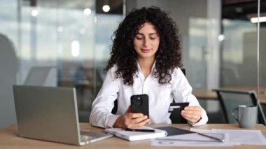 Kıvırcık saçlı, elinde kredi kartı ve akıllı telefonu olan gülümseyen bir iş kadını. Modern çalışma alanı, başarılı kadın yönetici, çevrimiçi bankacılık.
