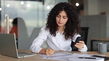 Odaklanmış iş kadını akıllı telefondaki verileri analiz eder ve notlar alır. Ofiste çalışan kıvırcık saçlı kadın. Modern iç mimari.