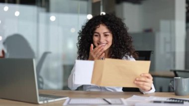 Heyecanlı iş kadını modern ofis masasında bir zarf açıyor. İçeride iyi haberler buluyor ve sevinçten havalara uçuyor..
