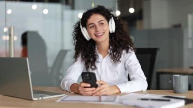 Gülümseyen iş kadını modern bir ofiste kulaklık ve telefon kullanarak müzik dinler. O, işte mola vermeyi sever..
