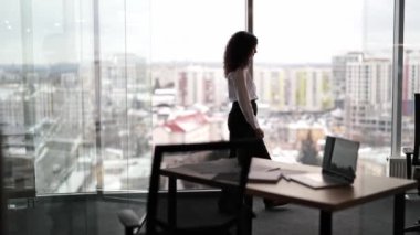 Kıvırcık saçlı düşünceli iş kadını modern ofiste duruyor ve şehir manzarasına geniş pencereden bakıyor..
