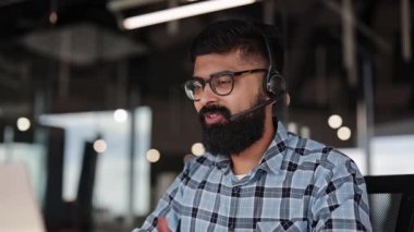 Sakallı ve gözlüklü gülümseyen işadamı kulaklıkla müşteriyle konuşuyor. Dost canlısı Hintli erkeğin modern ofiste video görüşmesi var. Kendine güvenen Hintli erkek yönetici, dizüstü bilgisayarla müşteriye danışmanlık yapıyor..