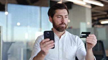 Akıllı telefon ve kredi kartı kullanan bir işadamı modern ofiste online ödeme yapıyor. Sakallı yönetici cep telefonuyla online alışveriş yapıyor.