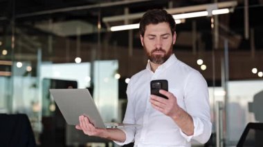 Modern ofiste dizüstü bilgisayar ve telefon kullanan odaklanmış bir iş adamı. Beyaz gömlekli sakallı adam iş projesinde çalışıyor..