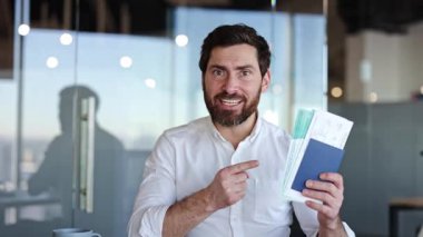 Heyecanlı iş adamı pasaportunu ve biniş kartını gösteriyor, iş gezisine hazır. Mutlu erkek profesyonel kameraya gülümsüyor, ofis masasında oturuyor..