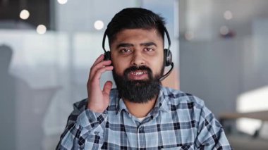 Mavi ekose gömlekli sakallı Hintli işadamı, sanal bir toplantı sırasında modern ofis ortamında kablosuz bir kulaklıkla konuşuyor..