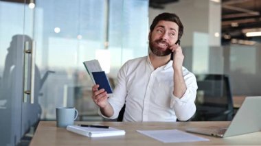 Sakallı ve beyaz gömlekli heyecanlı iş adamı pasaport ve biletleri tutuyor ve ofis masasında akıllı telefondan konuşuyor. O, yurt dışına seyahat etmekten mutlu..