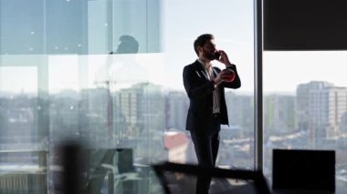 Takım elbiseli kendine güvenen işadamı, şehir manzarasına bakan pencerenin yanındaki modern ofiste top oynarken telefonla konuşuyor..