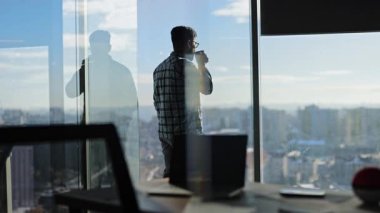 Genç işadamı şehir manzarasında modern bir ofis penceresinden dışarı bakarken sabah kahvesinin tadını çıkarıyor.
