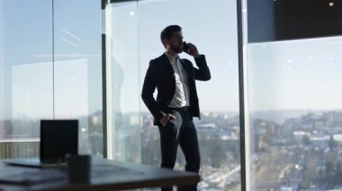 Modern ofiste şehir manzaralı kendine güvenen bir işadamı telefonda konuşuyor. Profesyonel adam iş anlaşmasını tartışıyor. Çağdaş çalışma alanındaki pencerenin yanında duruyor..