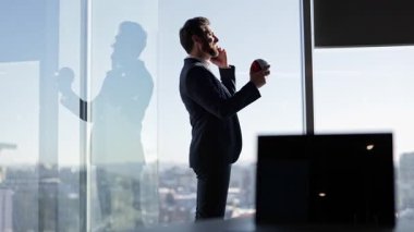 Kendine güvenen iş adamı telefonda konuşuyor ve modern gökdelendeki ofis pencerelerinde stres topuyla hokkabazlık yapıyor. Şehir manzarasını seviyor..