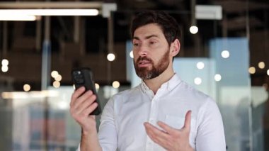 Kendine güvenen iş adamı, modern ofiste telefonla konuşuyor. Beyaz gömlek giyen ve görüntülü konuşma yapan genç bir erkek profesyonel..