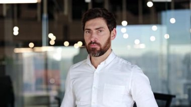 Çapraz kollu kendine güvenen işadamı modern ofiste duruyor ve gülümsüyor. Çağdaş çalışma alanında beyaz gömlekli sakallı bir adam..